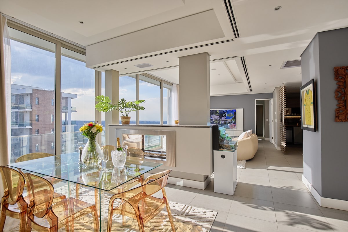A bright living area showcases a glass dining table surrounded by transparent chairs. Large windows allow natural light and offer views of the exterior. A cozy seating area is visible, complemented by a contemporary television mounted on a divider wall. A decorative vase holds fresh flowers.