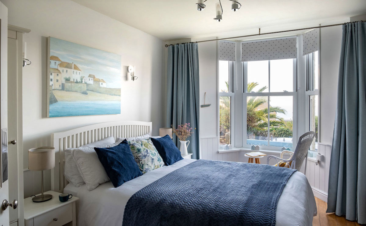 A bright bedroom features a spacious bed adorned with navy and floral cushions, and a lightweight blanket. Large windows allow natural light and showcase a view of greenery outside. Soft blue curtains frame the windows, while a beach-inspired painting complements the coastal decor.