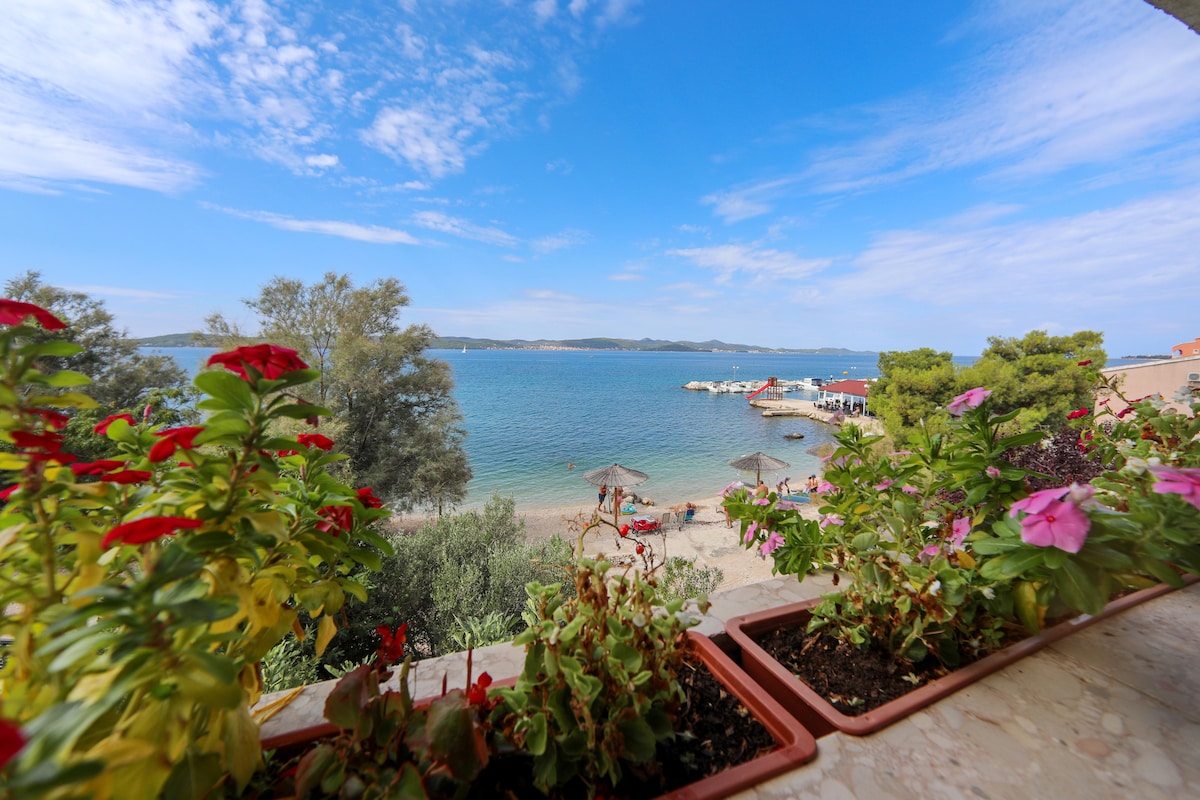 A view of the tranquil beach is framed by vibrant flower pots in the foreground. The clear blue water stretches towards the horizon, with a sandy shore visible. A few umbrellas and people can be seen relaxing by the water's edge, under a bright sky with scattered clouds.