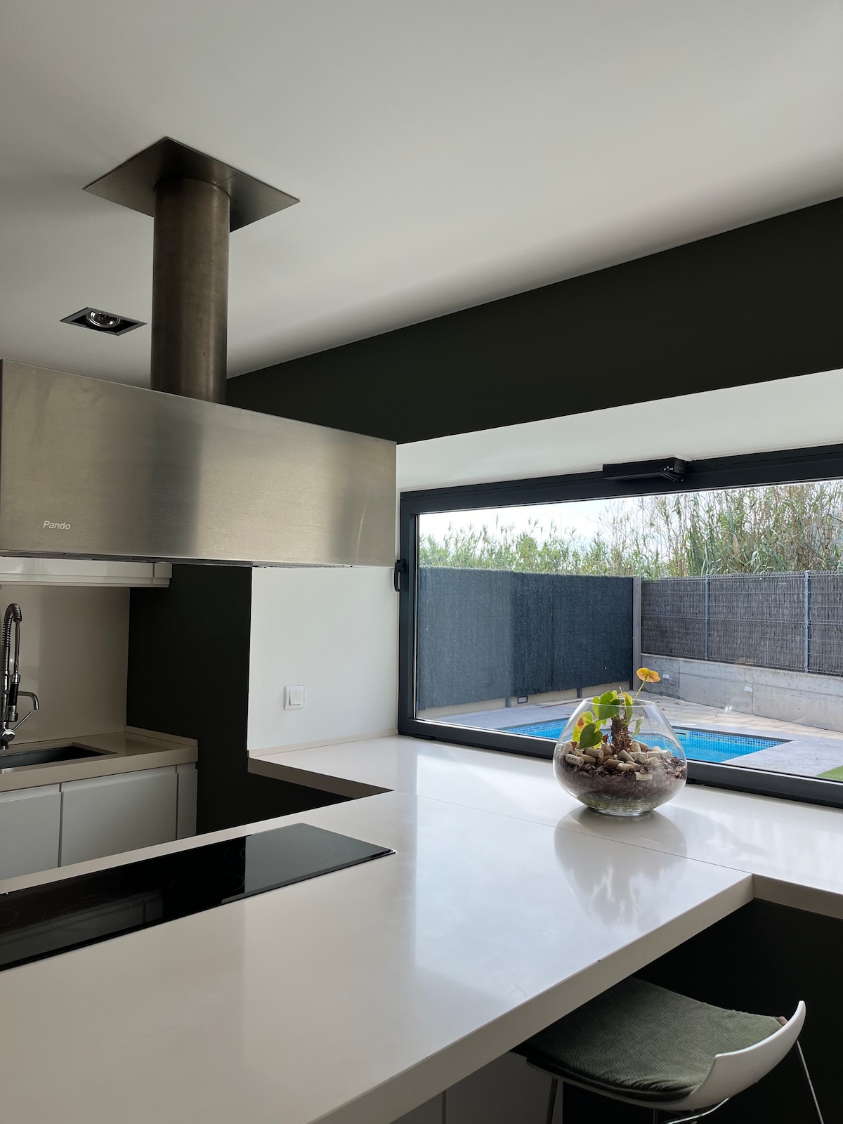 A modern kitchen features a sleek cooking area with a stainless steel hood above a smooth countertop. Large windows provide a view of the outdoor space and garden, with natural light enhancing the neutral color palette.