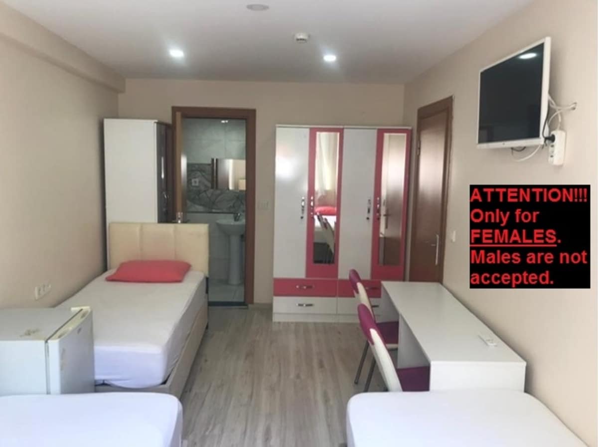 A spacious room is presented with two beds and a desk, featuring a flat-screen television mounted on the wall. Storage cabinets with mirrored doors provide additional space. A private bathroom is visible in the background. Clear indications of availability for female guests exclusively are noted.