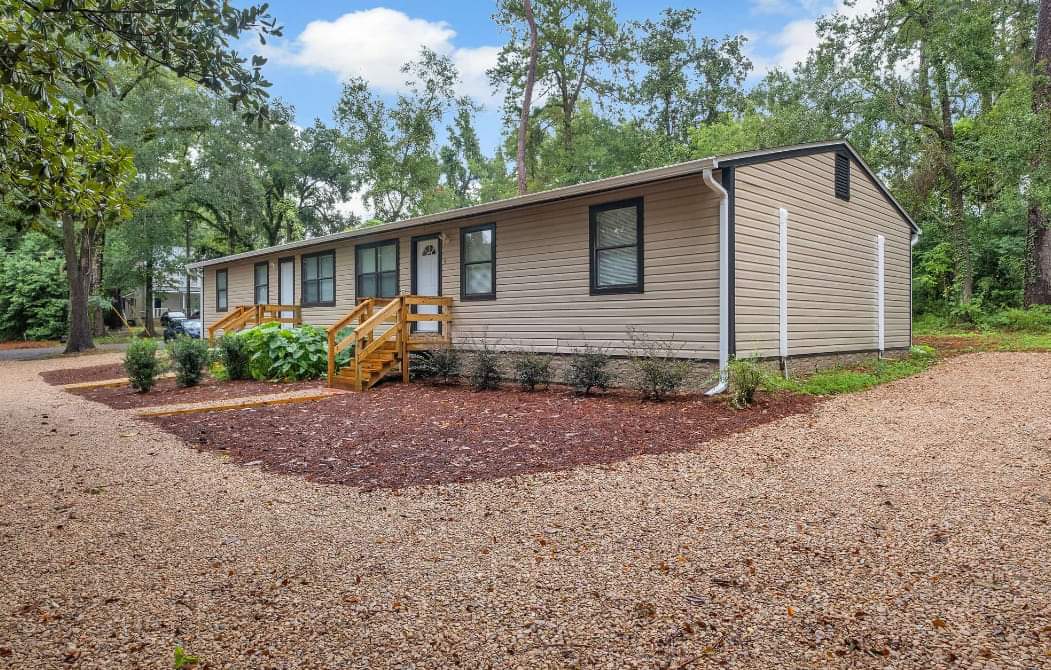 A single-story home is set among lush greenery, featuring a light-colored exterior. Two wooden steps lead up to the front door, complemented by manicured shrubbery along the pathway. A gravel driveway provides ample parking space, surrounded by trees that add to the serene setting.