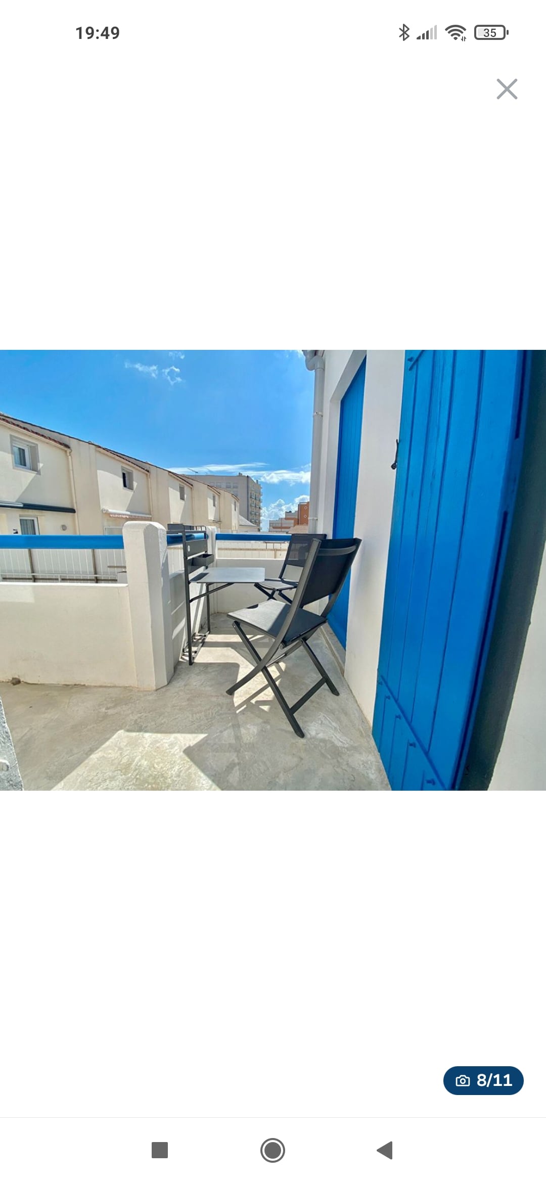 A balcony features two black folding chairs and a small table, creating a space for outdoor seating. The blue door provides a vibrant contrast against the light walls. A clear sky and distant buildings can be seen in the background, enhancing the area’s open feel.