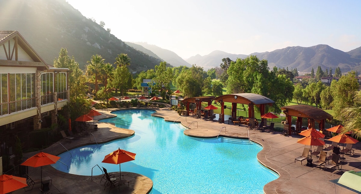 A large swimming pool is surrounded by vibrant orange umbrellas and lounge chairs, set against a backdrop of lush greenery and mountainous terrain. The sun casts a warm glow over the scene, highlighting the tranquil atmosphere of the resort.