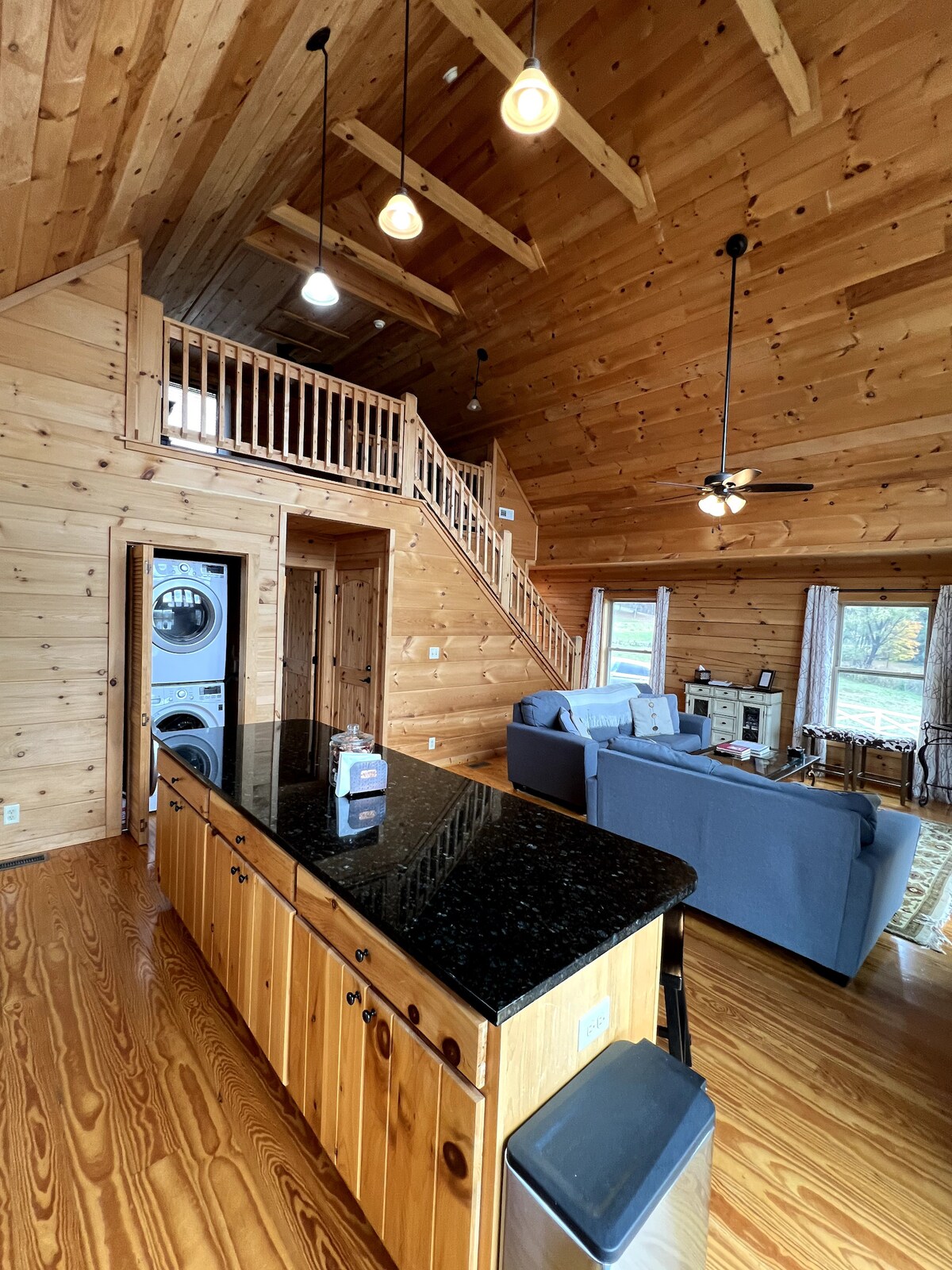 An open floor plan reveals a spacious living area featuring wooden walls and vaulted ceilings. A sleek granite countertop island separates the kitchen from the lounge area, with modern appliances visible. A staircase leads to a lofted area, and a washer and dryer are located in the corner.