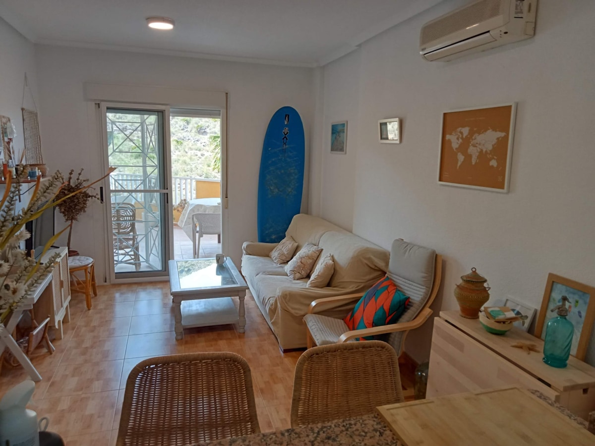 A living area features a comfortable sofa adorned with colorful cushions. A coffee table is placed in front of the sofa, while a surfboard leans against the wall. Large glass doors provide access to an outdoor space with additional seating.