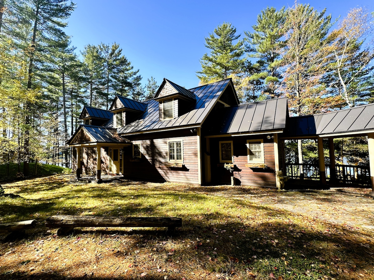 A spacious cabin is set against a backdrop of tall trees, showcasing a blend of dark wood siding and a metal roof. The exterior features multiple gables and several windows, with an outdoor area covered in leaves, indicating the beauty of the natural surroundings.