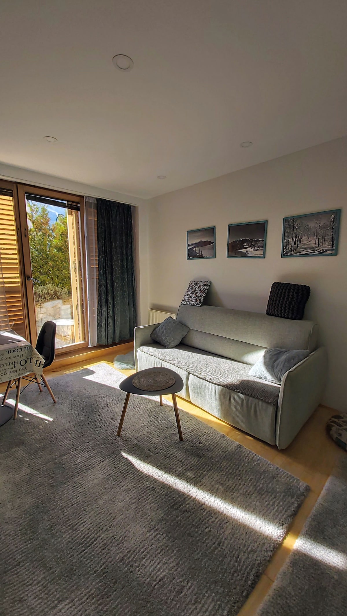 A comfortable sitting area is presented with a light-colored sofa adorned with cushions. A small round table is positioned nearby, and the space is illuminated by natural light coming through large sliding doors that lead to a terrace. Wall art adds character to the room.