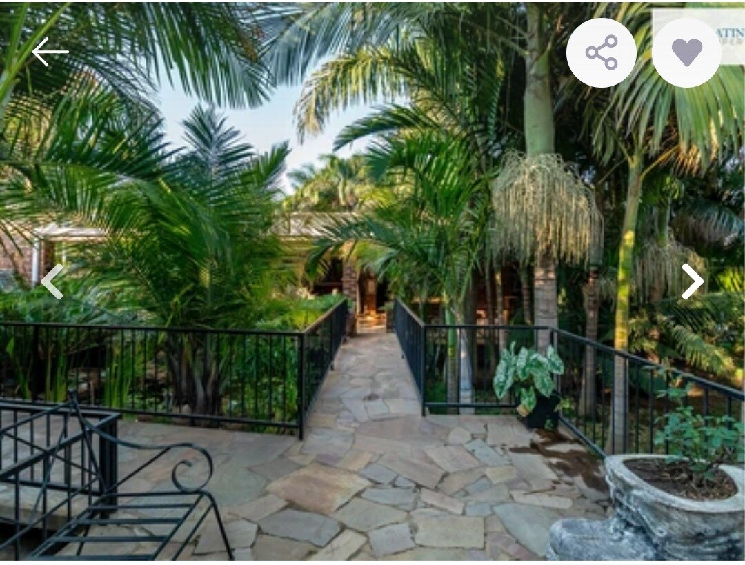 A pathway made of natural stone is bordered by lush palm trees and tropical foliage. Black wrought iron railings frame the walkway, which leads toward a shaded area in the distance, enhancing the serene garden atmosphere.