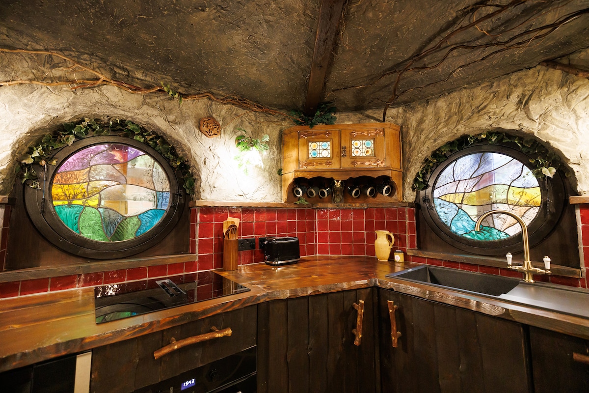 A unique kitchen features artistic stained glass windows with vibrant color patterns, set against textured walls. Wooden cabinetry and a countertop are complemented by modern kitchen appliances, including a stovetop and sink, creating a cozy yet functional space.