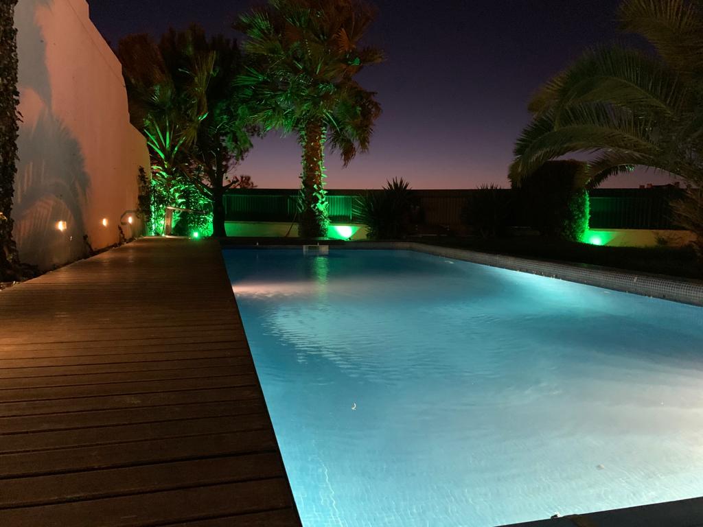 A swimming pool is illuminated with soft green lights, surrounded by palm trees and colorful landscaping. A wooden deck extends alongside the water, creating a tranquil outdoor space. The evening sky casts a subtle gradient, enhancing the peaceful atmosphere.