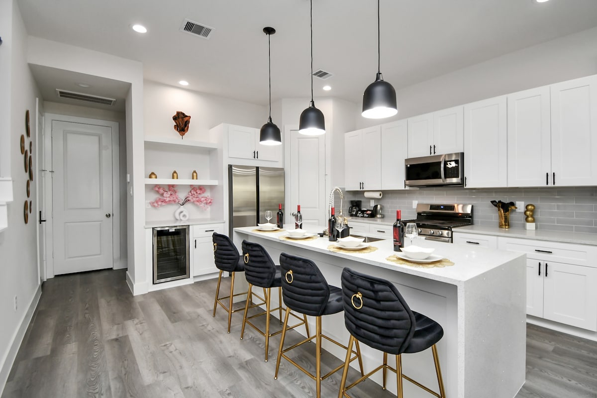 A modern kitchen is shown, featuring stainless steel appliances, including a refrigerator and a built-in wine cooler. A large island with four black bar stools is centered, with dinnerware arranged neatly. Pendant lights hang overhead, illuminating the space with a sleek ambiance.
