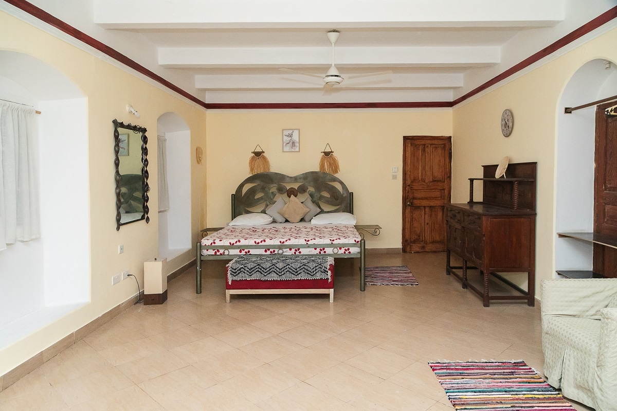 A spacious bedroom features a large bed adorned with neutral-toned linens and decorative pillows. Natural light enters through a window dressed with sheer curtains. The room is complemented by a wooden dresser and a wall mirror, with traditional rugs adding warmth to the tiled floor.