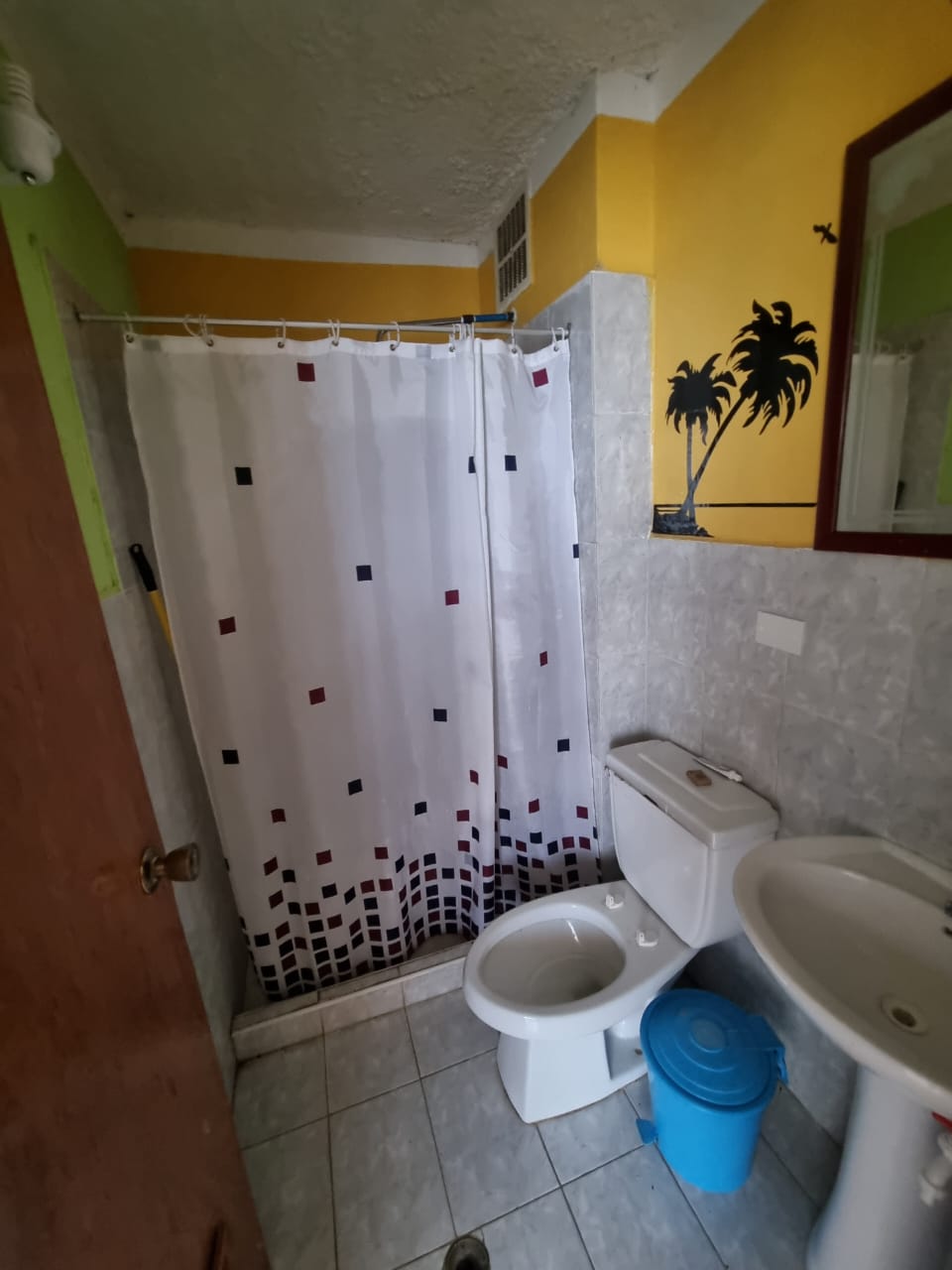 The bathroom features tiled flooring and walls, with a shower area enclosed by a patterned curtain. A toilet and sink are positioned against the wall, complemented by a small mirror above the sink. A blue bucket is visible on the floor.