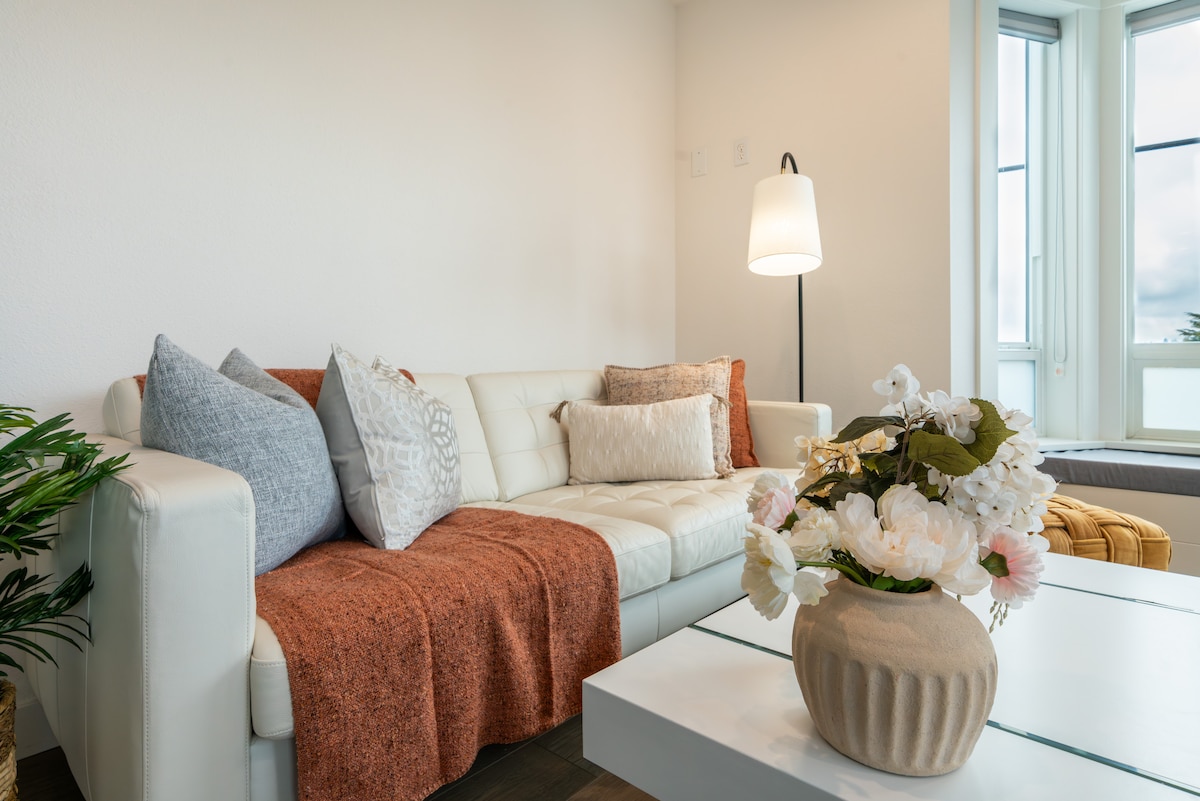 A light-colored sofa adorned with various textured pillows is positioned near a table topped with a simple vase of flowers. A lamp stands beside the sofa, and large windows provide natural light, enhancing the room's openness and view.
