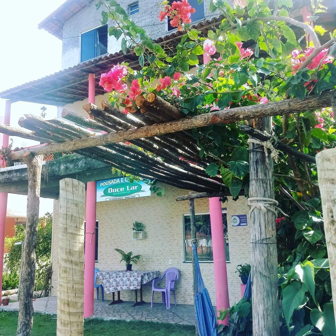 A charming exterior view features a shaded pergola adorned with vibrant flowers. The entrance to the pousada is visible, showcasing a welcoming sign. A dining table and chairs are set under the pergola, surrounded by lush greenery, creating an inviting atmosphere.