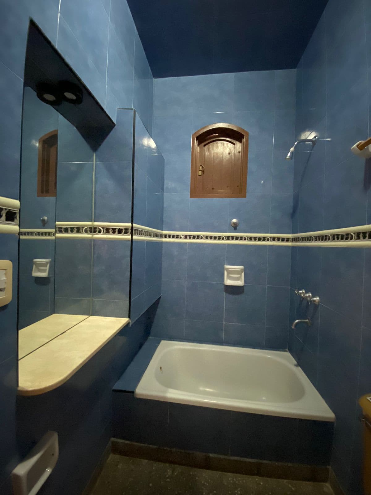 A bathroom featuring blue tiled walls and a bathtub with a shower fixture. A small wooden window is set above the bathtub, and a marble countertop with a mirror extends alongside the wall. The bathroom is equipped with essential toiletries and fixtures.