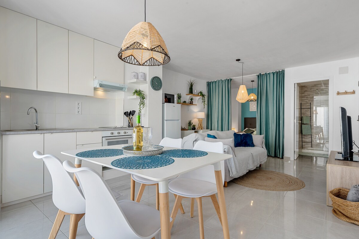 A contemporary open-plan living area is presented, featuring a dining table with four chairs and decorative placemats. A well-equipped kitchen is visible, alongside a cozy living space with a light-colored sofa and soft blue décor. Lantern-style light fixtures enhance the overall ambiance.
