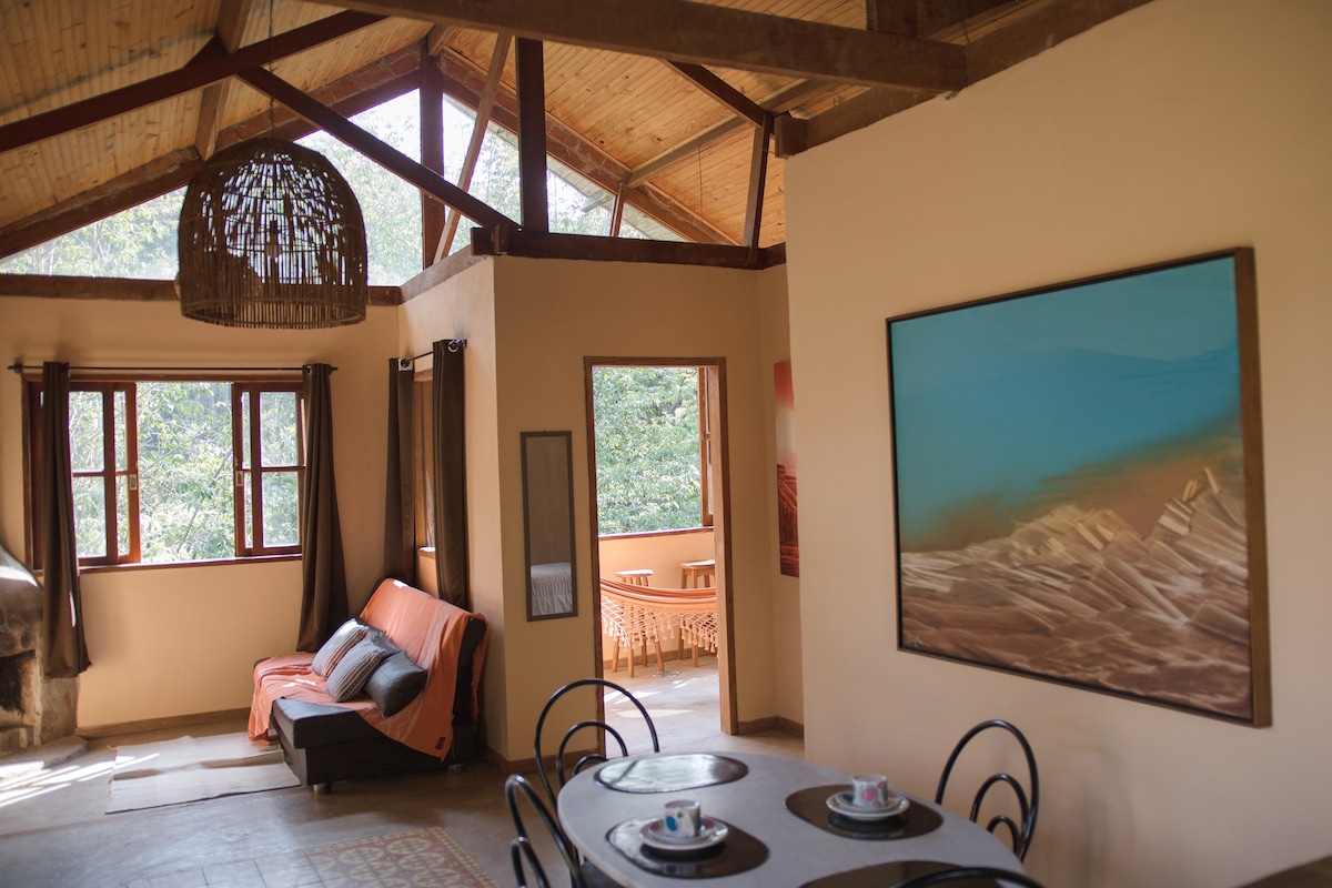 A spacious living area features large windows that bring in natural light, showcasing views of the surrounding greenery. A dining table is set for four, with decorative plates and cups. A cozy sofa sits nearby, accompanied by earthy tones and wooden beams overhead.