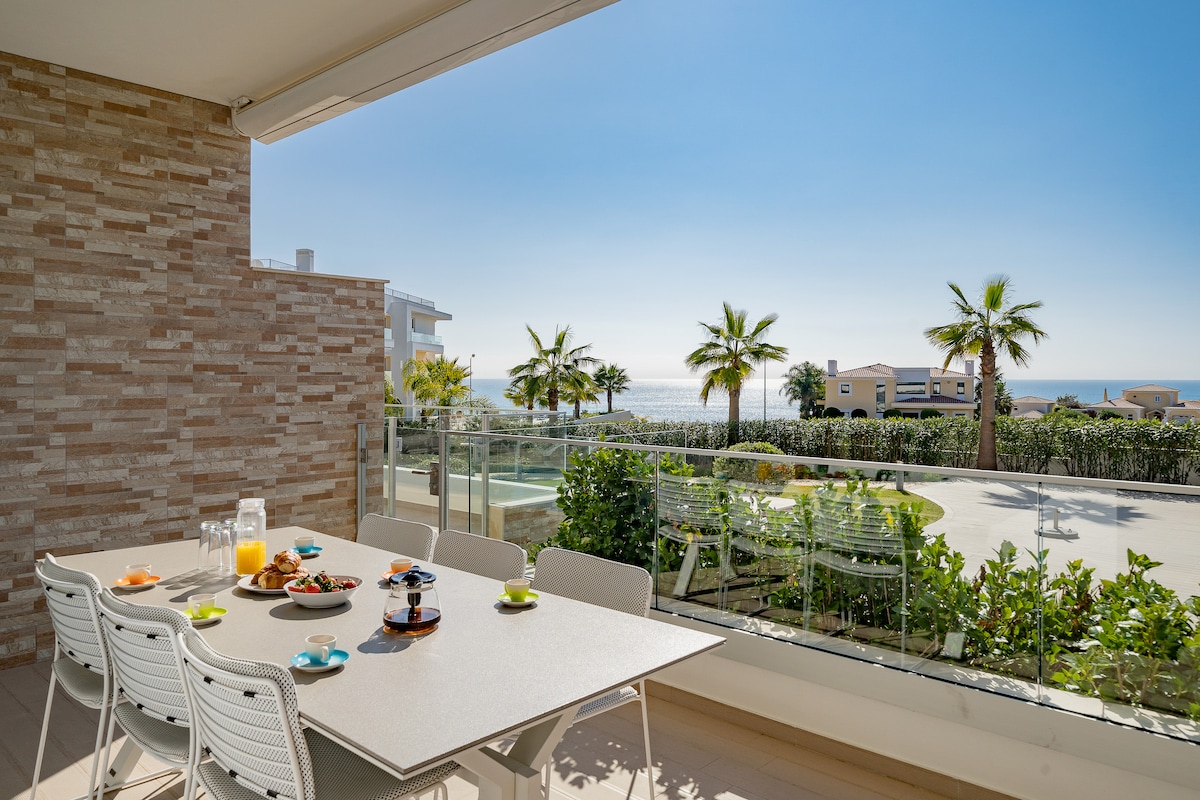 A spacious terrace is featured, equipped with a dining table surrounded by white chairs. The view showcases palm trees and the sparkling ocean in the distance, with well-maintained greenery and a communal pool visible below.