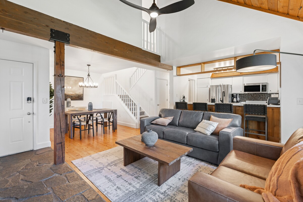 The living area features a mix of textures with a leather sofa and a fabric couch facing a wooden coffee table. A large rug anchors the space, while light streams in from windows above. The open layout connects to the dining area and modern kitchen.