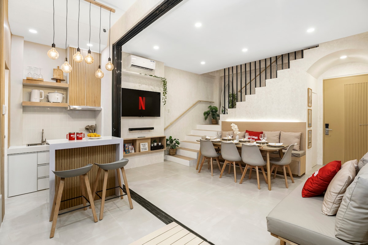 A modern living space features a kitchenette with bar seating for two, a dining area with a long table surrounded by six chairs, and a cozy lounge area. Soft lighting and a wall-mounted TV with Netflix create a welcoming environment, enhanced by natural materials and textures.