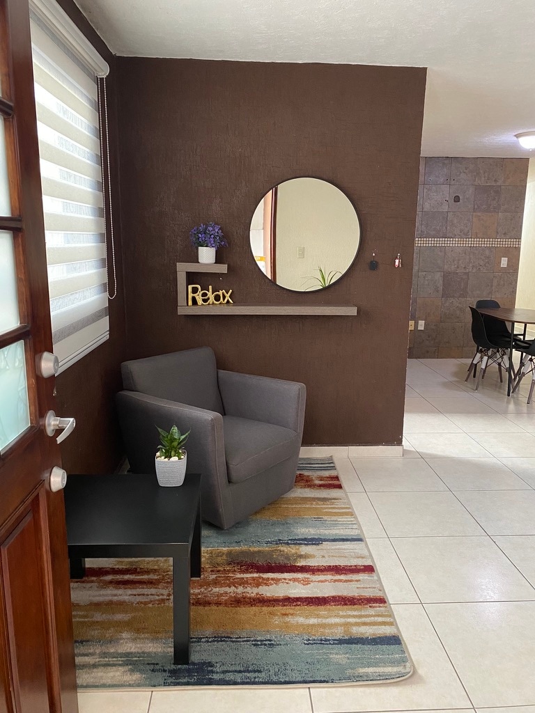 A welcoming entryway presents a cozy gray armchair beside a small black table with a plant. A decorative shelf displays a small potted plant and the word 'Relax.' The wall features a circular mirror, enhancing the space’s brightness and inviting atmosphere.