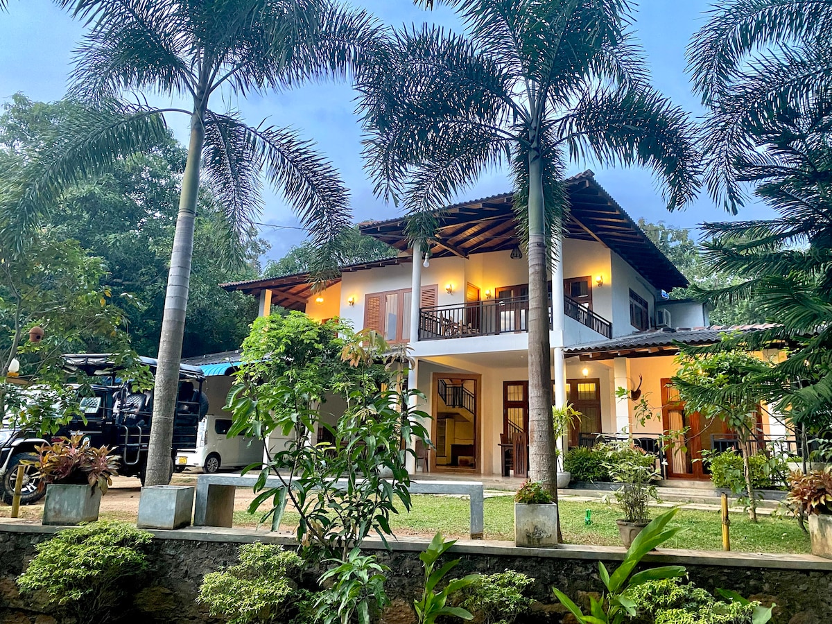 A two-story house with a welcoming entrance is surrounded by lush greenery and palm trees. The architecture features covered balconies and warm lighting, creating a cozy ambiance. A spacious yard showcases various plants, enhancing the tranquil outdoor setting.