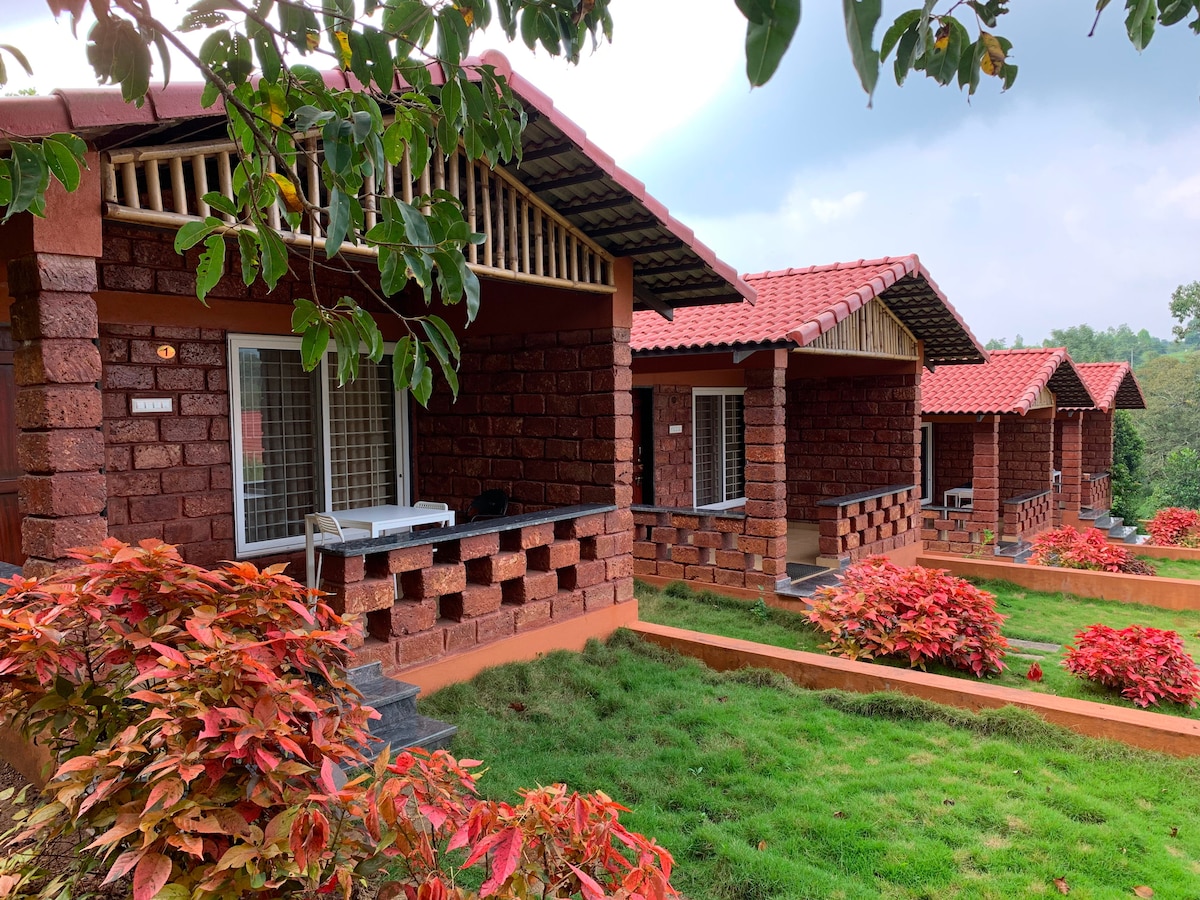 Four charming cottages with stone exteriors are featured, surrounded by vibrant red foliage. Each cottage has a spacious porch area with a small table and chairs, blending harmoniously with the lush green landscape and offering a peaceful retreat.