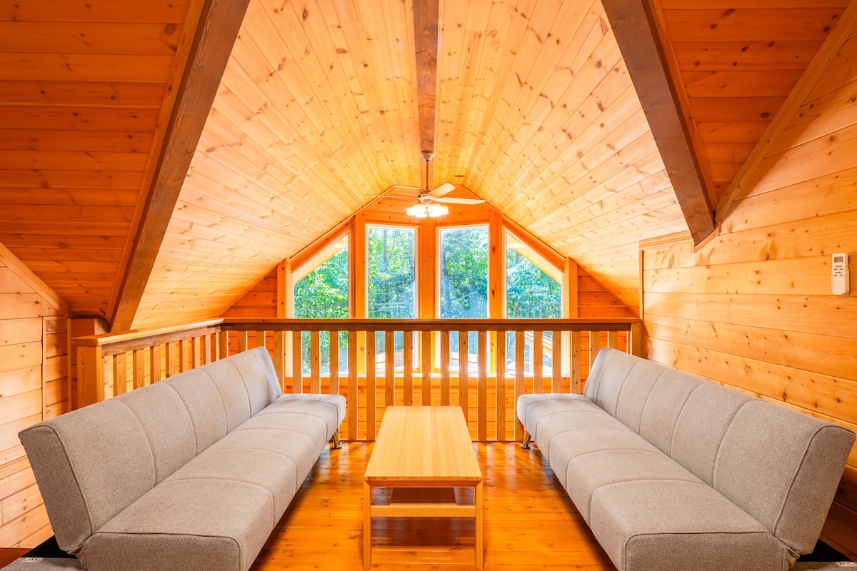 An inviting loft area is shown, featuring two spacious sofas arranged symmetrically around a wooden coffee table. Large windows provide ample natural light and offer scenic views of the surrounding greenery. The wooden walls and ceiling create a warm atmosphere.