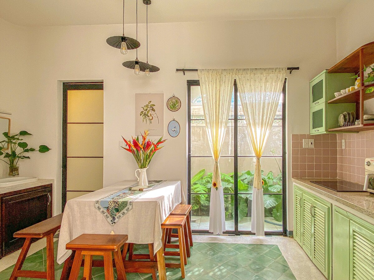 A bright kitchen features a wooden table surrounded by three stools, adorned with a centerpiece of tropical flowers. Natural light streams through large glass doors, providing views of lush greenery outside. Cabinets in soft green contrast with the warm tones of the countertop.