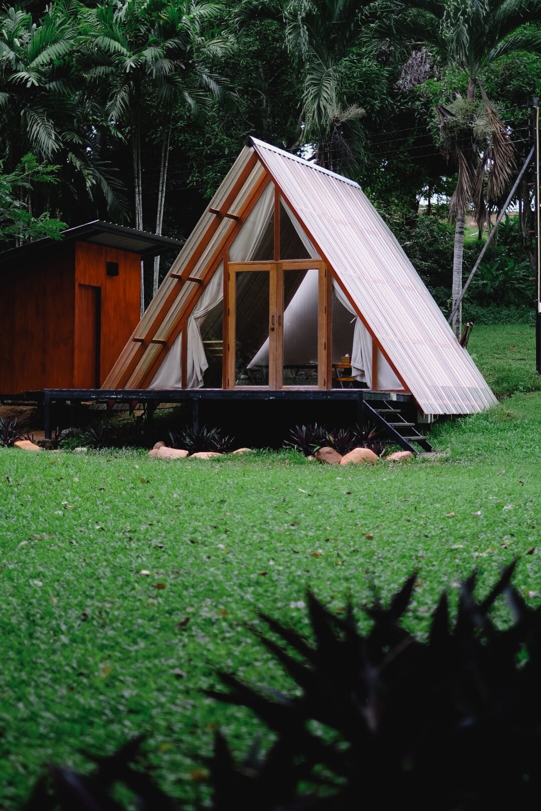 A triangular-shaped cabin with wooden framing and a sloped roof is set on a grassy area, surrounded by lush greenery. The front features large glass doors, inviting natural light into the interior. A small building can be seen adjacent to the cabin.