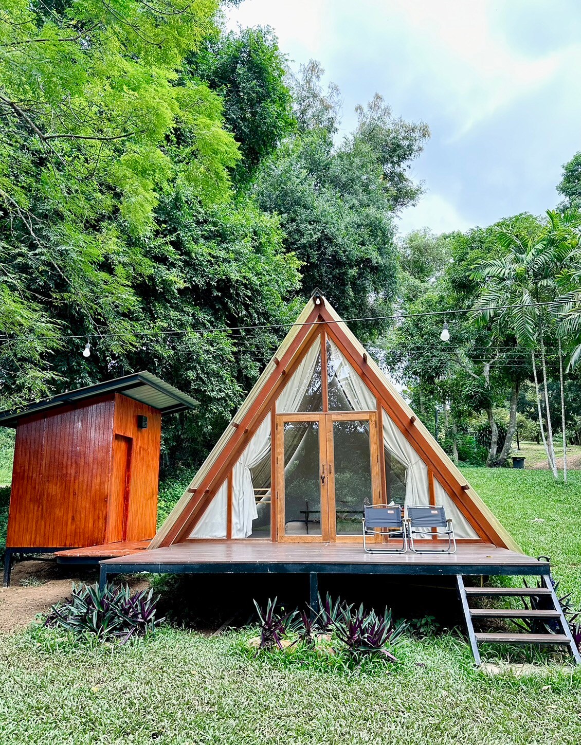A triangular wooden cabin is surrounded by lush greenery. The cabin features a large glass door at its front, leading to an outdoor area with two chairs. A small utility building stands nearby, contrasting with the natural surroundings.