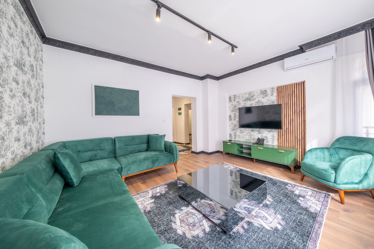The spacious living room features a green sofa set arranged around a low coffee table, situated on a patterned area rug. A wall-mounted television is positioned across from the seating area, and natural light enters through large windows, enhancing the room's openness.
