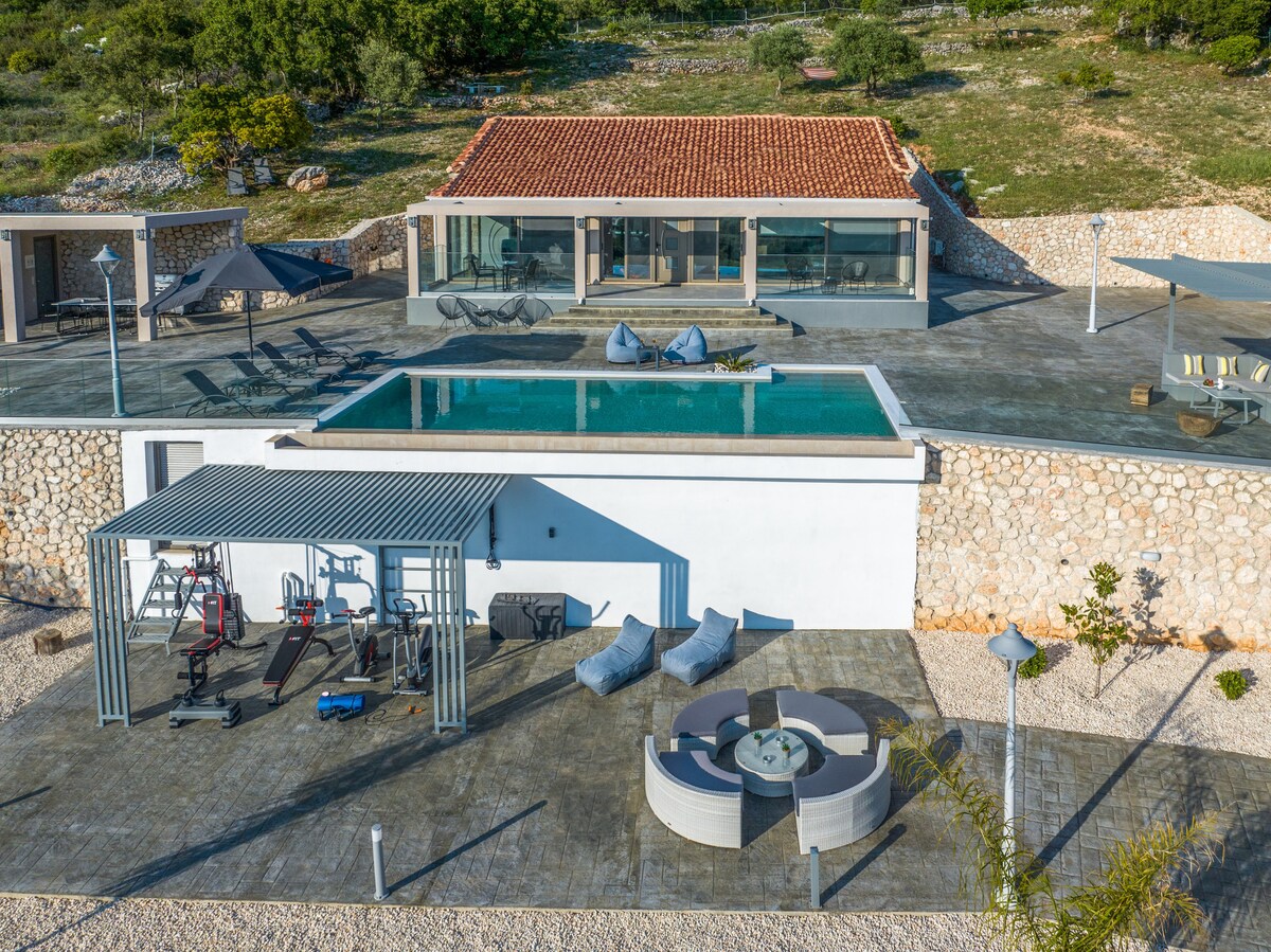 An aerial view of the villa reveals a spacious terrace featuring an infinity pool surrounded by sun loungers. An outdoor gym is visible nearby, equipped with various exercise machines. A shaded sitting area with a fire pit complements the outdoor space.