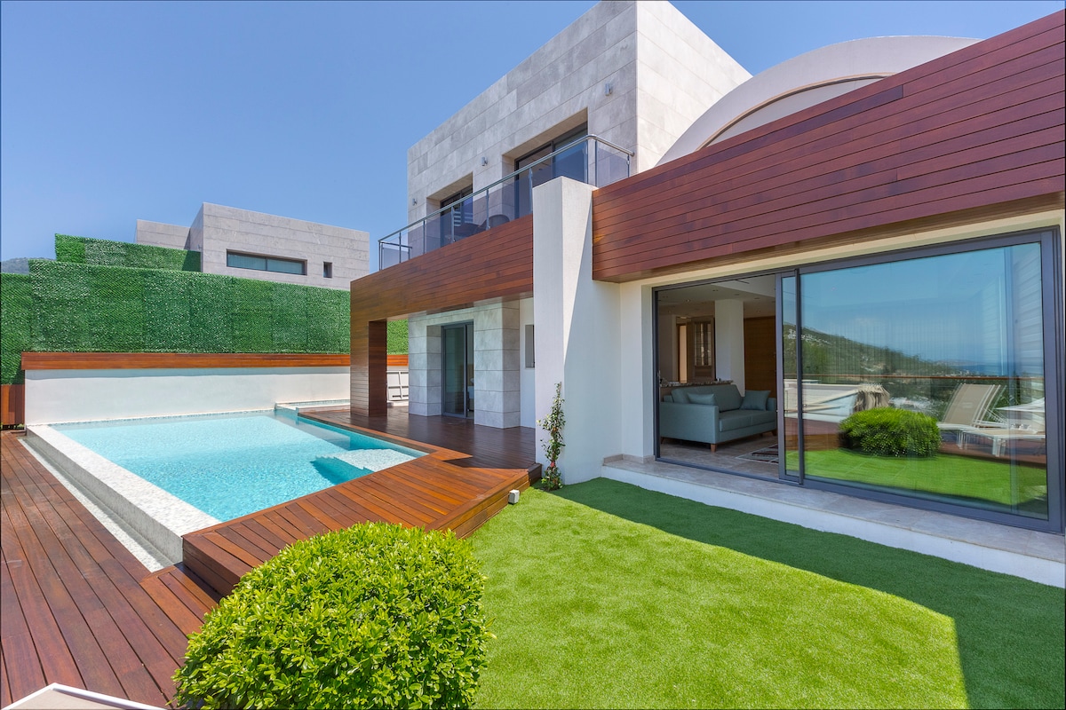 An inviting outdoor space features a private swimming pool surrounded by wooden decking and manicured green grass. The modern villa displays a blend of stone and wood elements, with large glass doors allowing natural light into the interior living area.