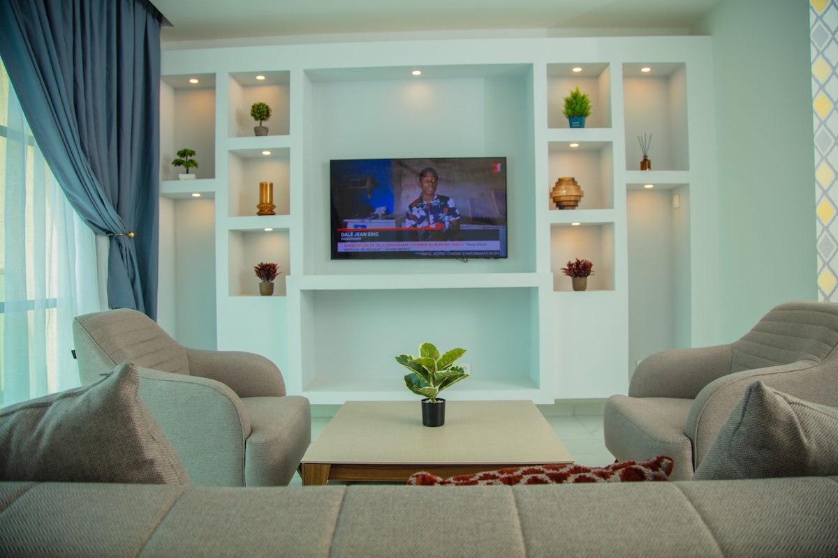 A modern living area is displayed, featuring a comfortable seating arrangement with light gray chairs and a sofa. A sleek television is mounted on a white wall unit, which is accented by decorative plants and soft lighting. Natural light filters in through large windows covered with elegant drapes.