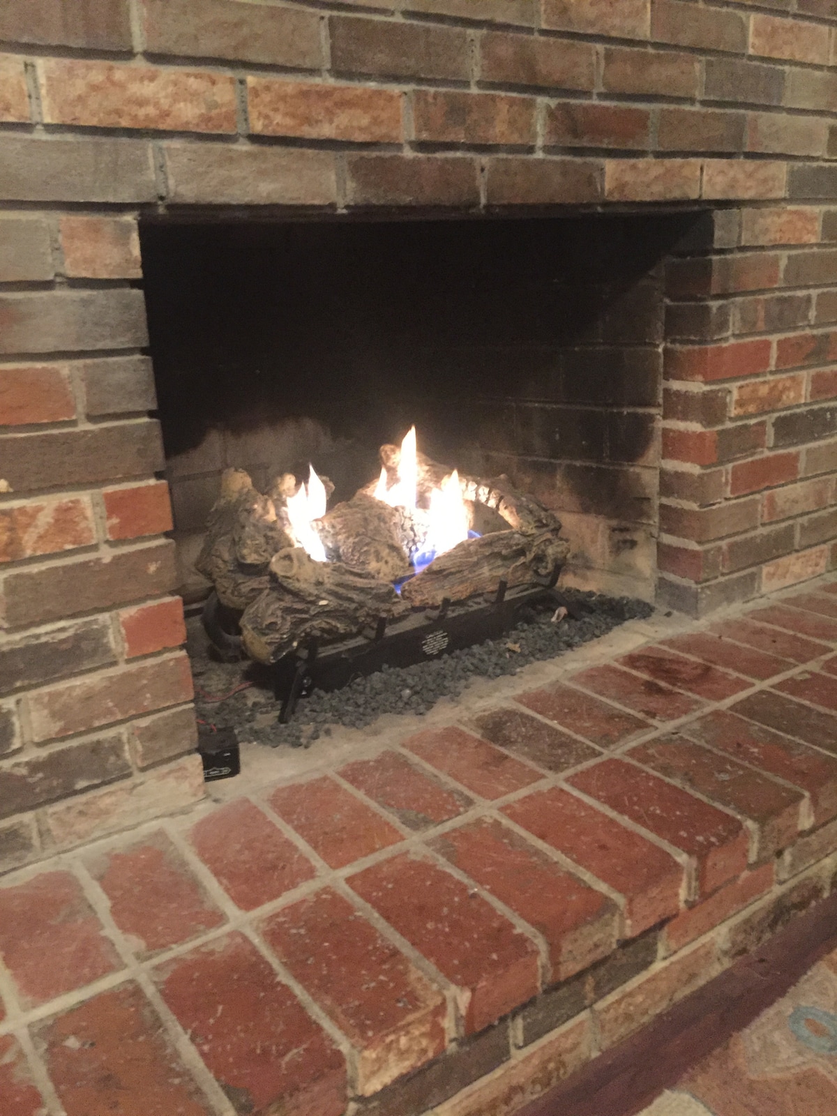 A traditional brick fireplace features a cozy flame, providing warmth to the surrounding space. The hearth is lined with red bricks, and the logs are arranged artfully within the firebox, creating a welcoming ambiance.