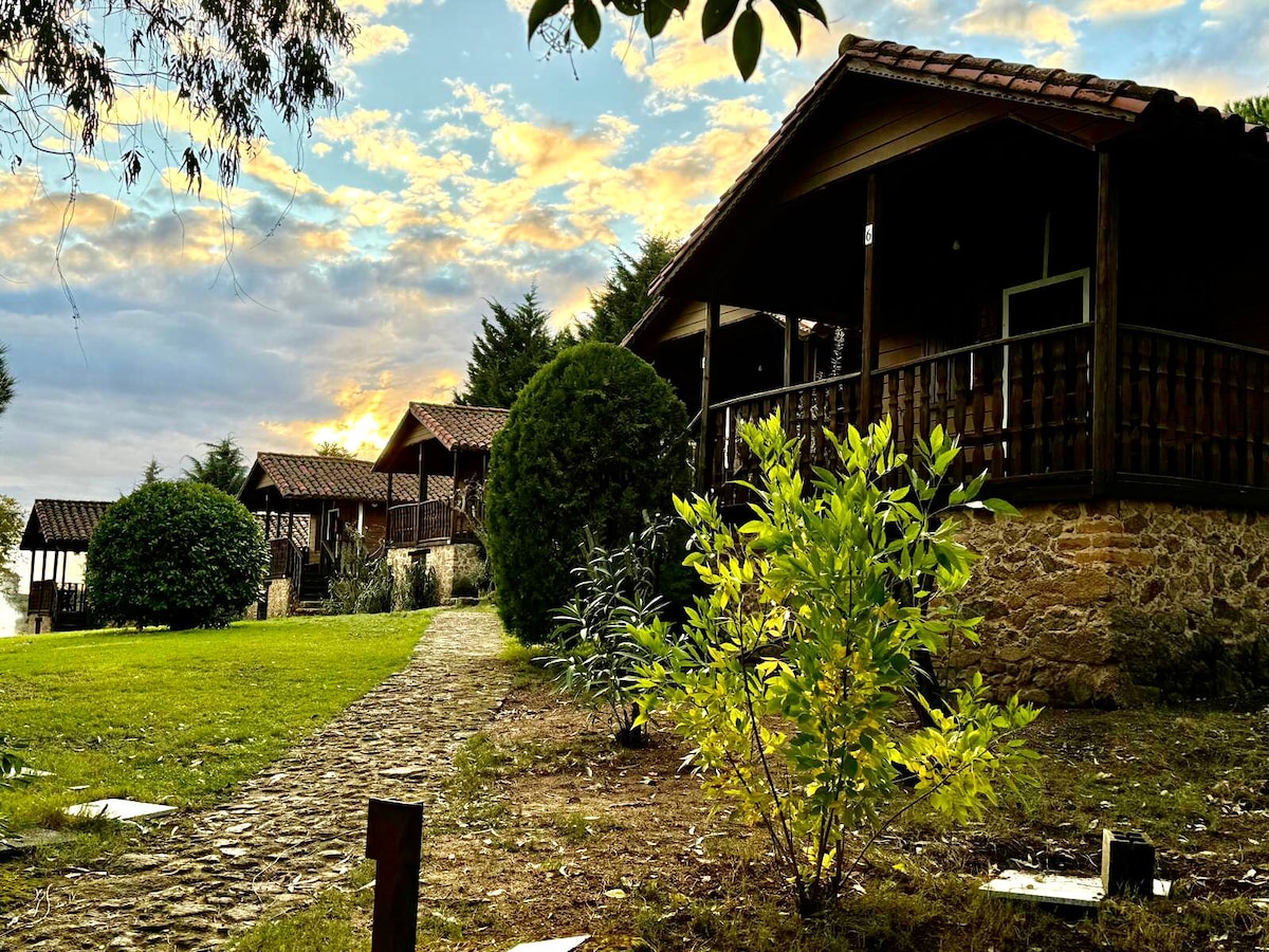 Wooden bungalows are nestled within a lush green landscape, surrounded by manicured gardens and trees. A winding stone pathway leads through the area, connecting the bungalows. The warm glow of sunset can be seen in the background, enhancing the serene environment.