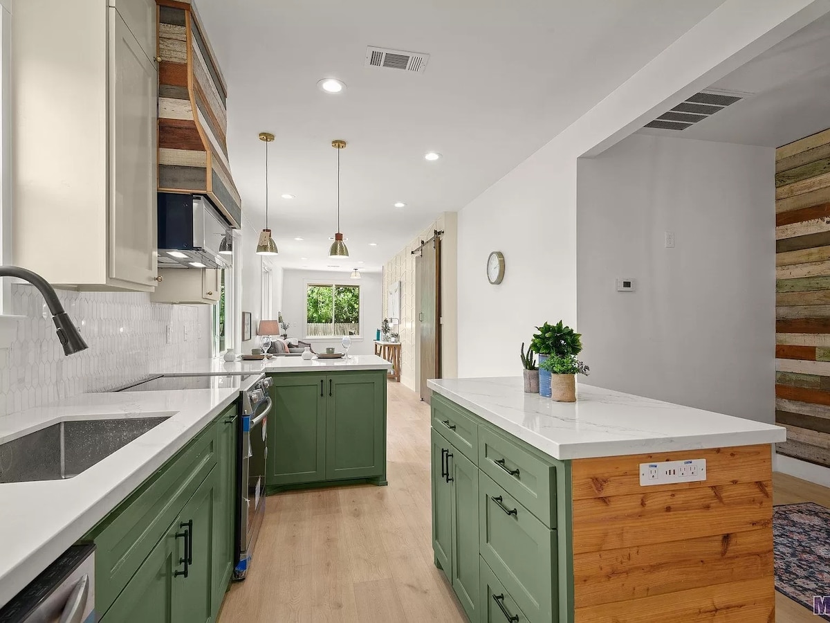 A modern kitchen features green cabinetry and white countertops. Pendant lights hang above the island, with a stainless steel sink and appliances visible. The space flows into a bright living area, complemented by natural light from large windows.