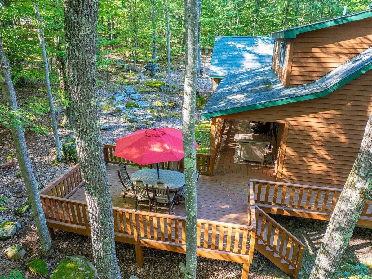 A spacious wooden deck is surrounded by trees, featuring a round dining table with chairs beneath a bright red umbrella. The natural landscape includes rocks and foliage, providing a serene outdoor setting. The deck connects to a warm-toned lodge with a sloping roof.