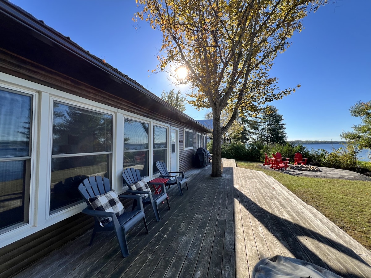A spacious outdoor deck is shown, featuring comfortable seating options facing the serene lake. Sunlight streams through the trees, creating a bright atmosphere. Red Adirondack chairs are arranged on the grassy area beneath the trees, offering inviting spots to relax and take in the view.