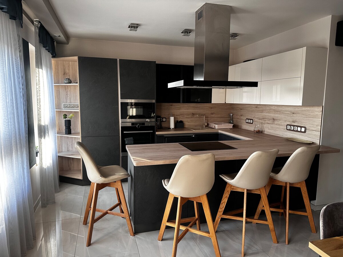 A modern kitchen features a large central island with four high-backed stools. Sleek cabinetry in contrasting colors is complemented by a stainless steel hood above the cooking area. Natural light enters through large windows, enhancing the contemporary design.