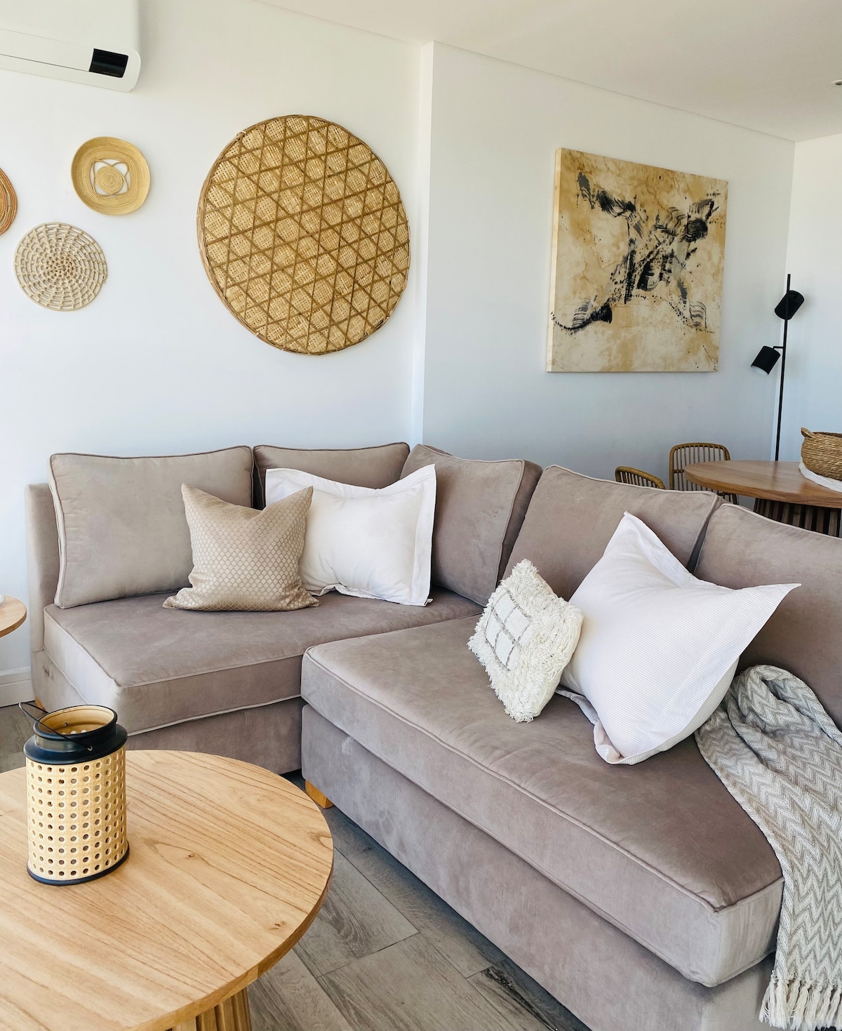 A soft sectional sofa in a neutral tone is surrounded by decorative wall hangings, including woven circular pieces. A round wooden table with a lantern adds warmth to the space, while a cozy throw blanket rests casually on the arm of the sofa.