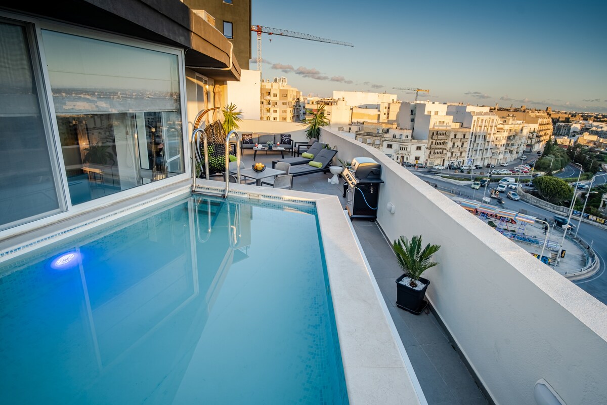 A rooftop terrace features a heated pool with a calm blue surface. Comfortable seating areas and sun loungers are arranged around the terrace, complemented by potted plants. The scenic view of the cityscape and distant skyline is visible, with construction cranes in the background.