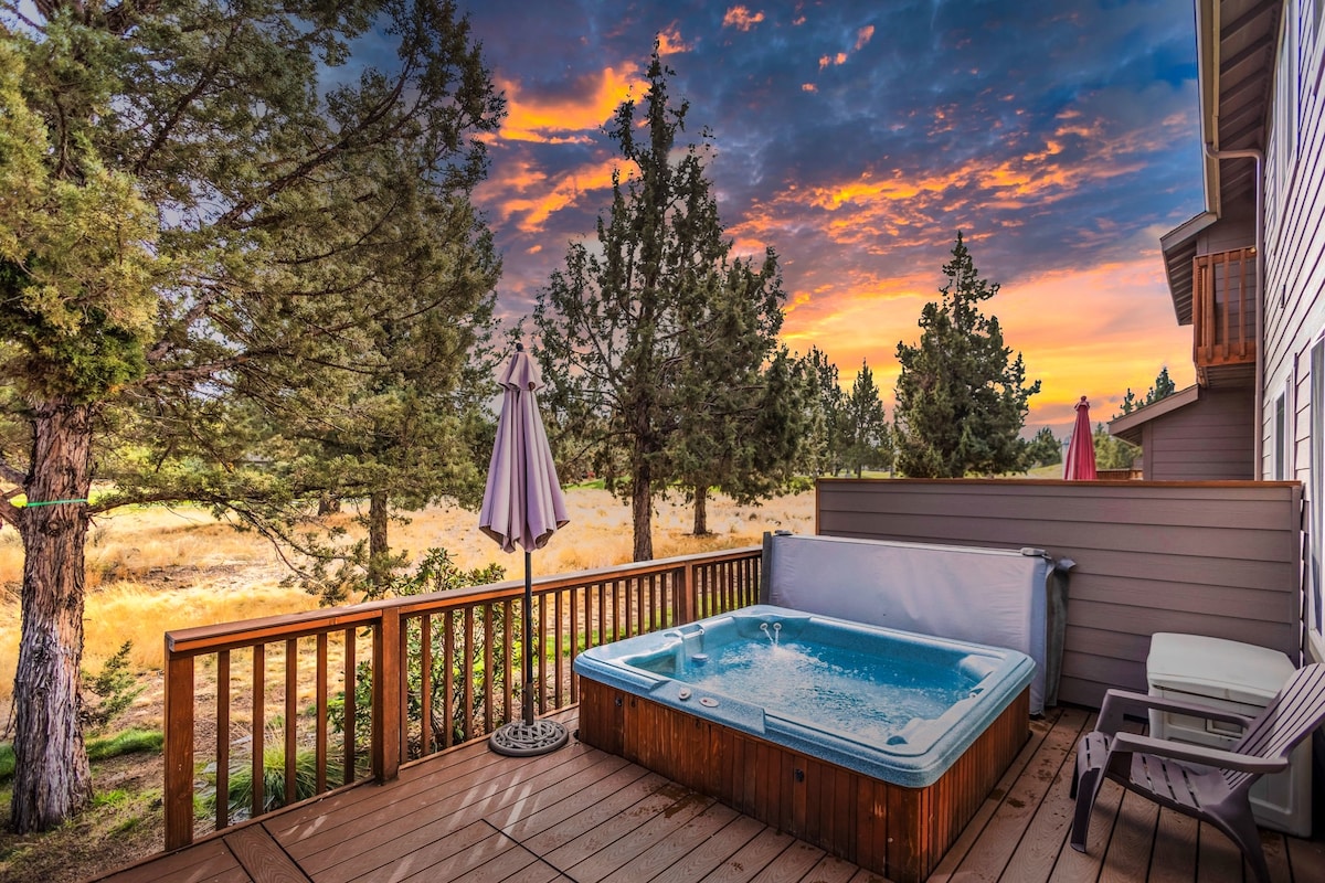 A private back deck features a hot tub, surrounded by trees with views of the golf course. A patio umbrella provides shade over a seating area, while the sky reflects vibrant colors during sunset. The wooden deck is inviting and spacious for relaxation.