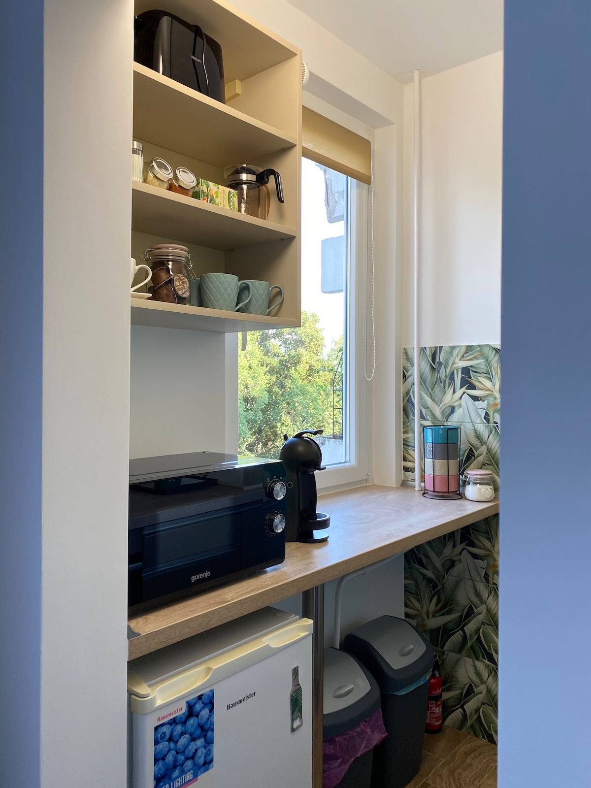 A compact kitchenette is visible, equipped with a microwave, coffee maker, and a refrigerator. Upper shelves hold various mugs and jars, while light streams in from a nearby window, illuminating a decorative wall with leafy patterns. Two trash bins are neatly placed beside the refrigerator.