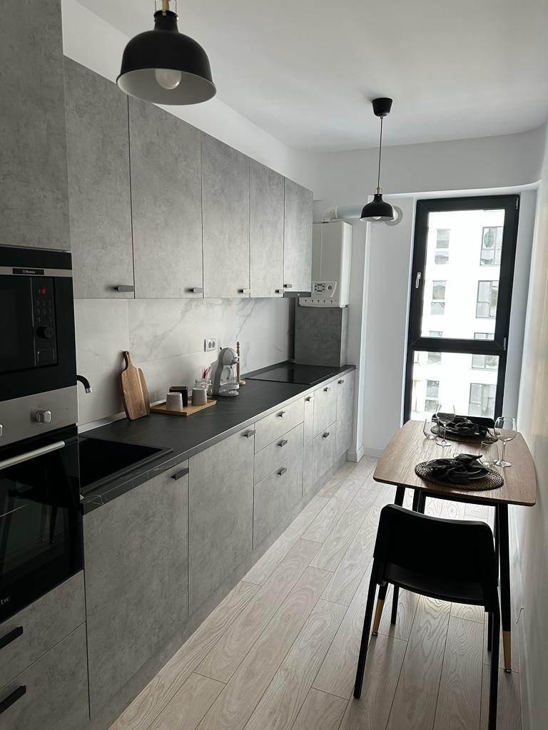A modern kitchen features sleek gray cabinets and a black countertop. An electric stove, oven, and refrigerator are included, along with a coffee maker and kettle. A small dining table with two chairs is set near a large window, allowing ample natural light.