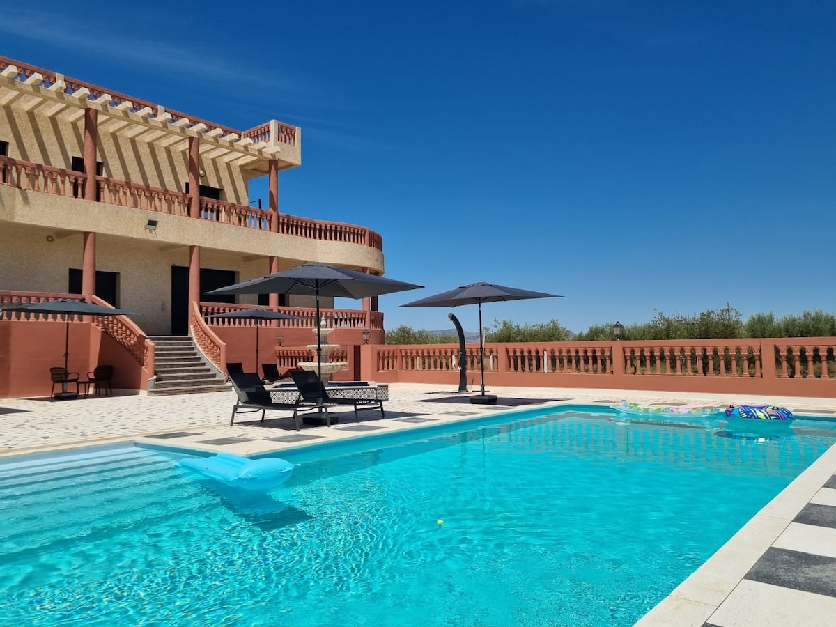 A spacious outdoor area features a large swimming pool surrounded by comfortable lounge chairs. Umbrellas provide shade, and the two-story villa is visible in the background, showcasing its architectural design against a clear blue sky.