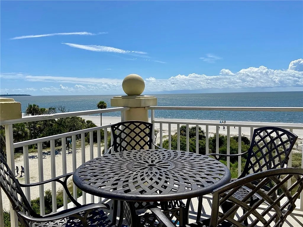 A round patio table with intricately designed metalwork is positioned on a balcony. The ocean stretches out in the background, with the sandy beach visible below. Lush greenery surrounds the area, and clear blue skies create a serene atmosphere.